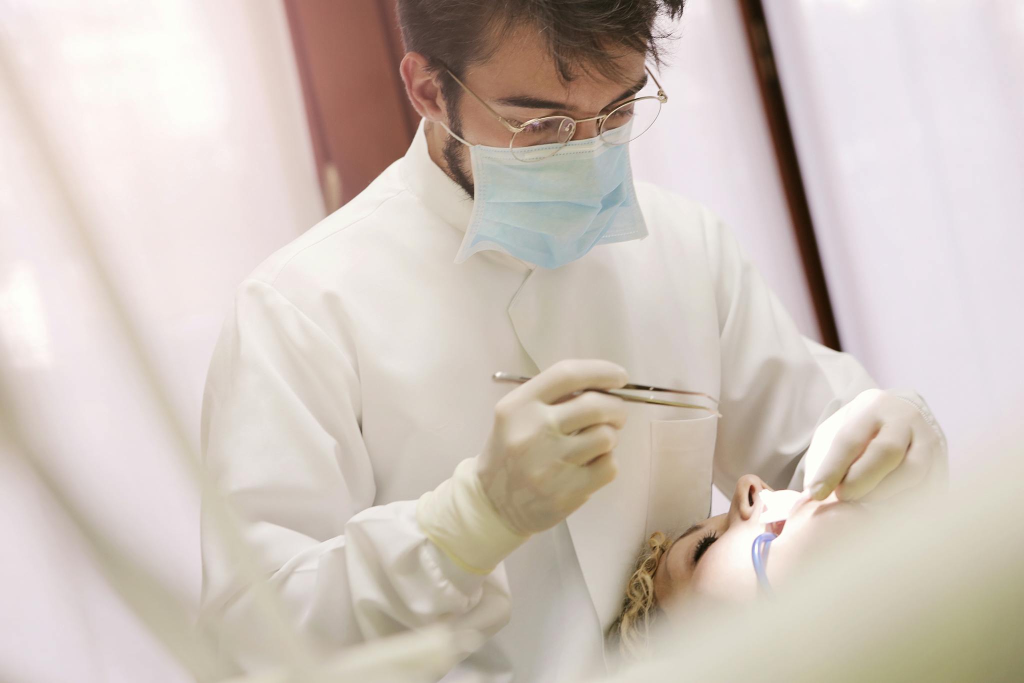 A dentist wearing a mask and gloves treating a patient at a dental clinic indoors.